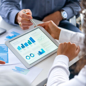 Close up of three people looking at financial data with graphs and charts. All their hands can be seen and one person is pointing with a pen. There is paperwork on the desk showing more finance information