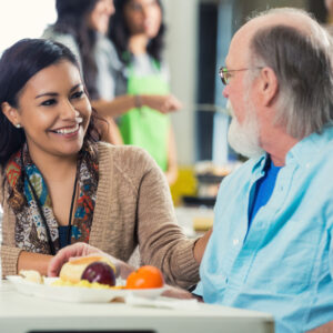 Young adult Hispanic woman is sitting at table in food bank or soup kitchen. She is smiling and talking to a senior Caucasian man. Man is enjoying a healthy meal. Volunteers are serving food to seniors in background.