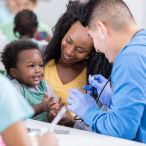 An adorable little toddler boy sits in his mother's lap in a volunteer run medical clinic and claps for a male nurse. He is unrecognizable as she leans in to listen to his heart.